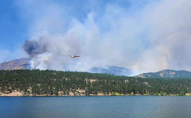 Wildlife smoke rises skyward from forested hills as a rescue helicopter flies toward it over a body of water