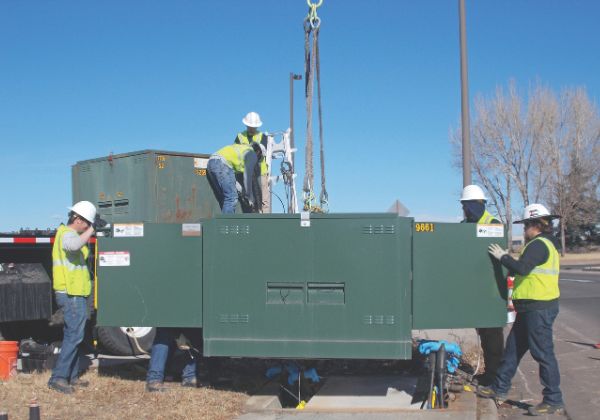 Utility workers in safety gear installing a large green electrical unit outdoors.