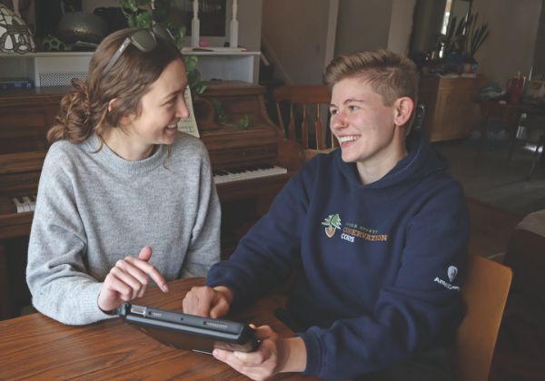 Two people sitting at a table smiling and looking at each other while one holds a tablet.